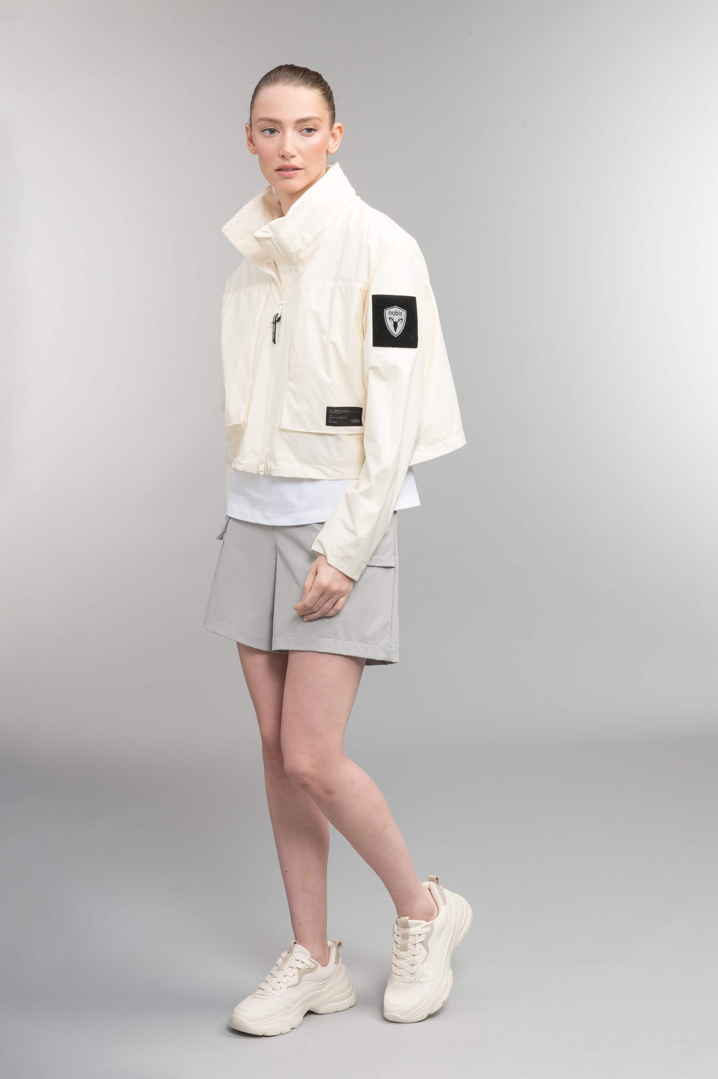 Epsie Women's Cropped Rain Jacket in Chalk crafted from premium recycled polyester ripstop with a DWR coating and taffeta nylon lining. Features a hidden packaway hood stored in the collar, two-way waterproof front zipper, seam-sealed construction, underarm zip ventilation, adjustable drawcord hem, zippered waist patch pockets, and an interior chest pocket for secure storage. Machine washable. Designed in Canada.