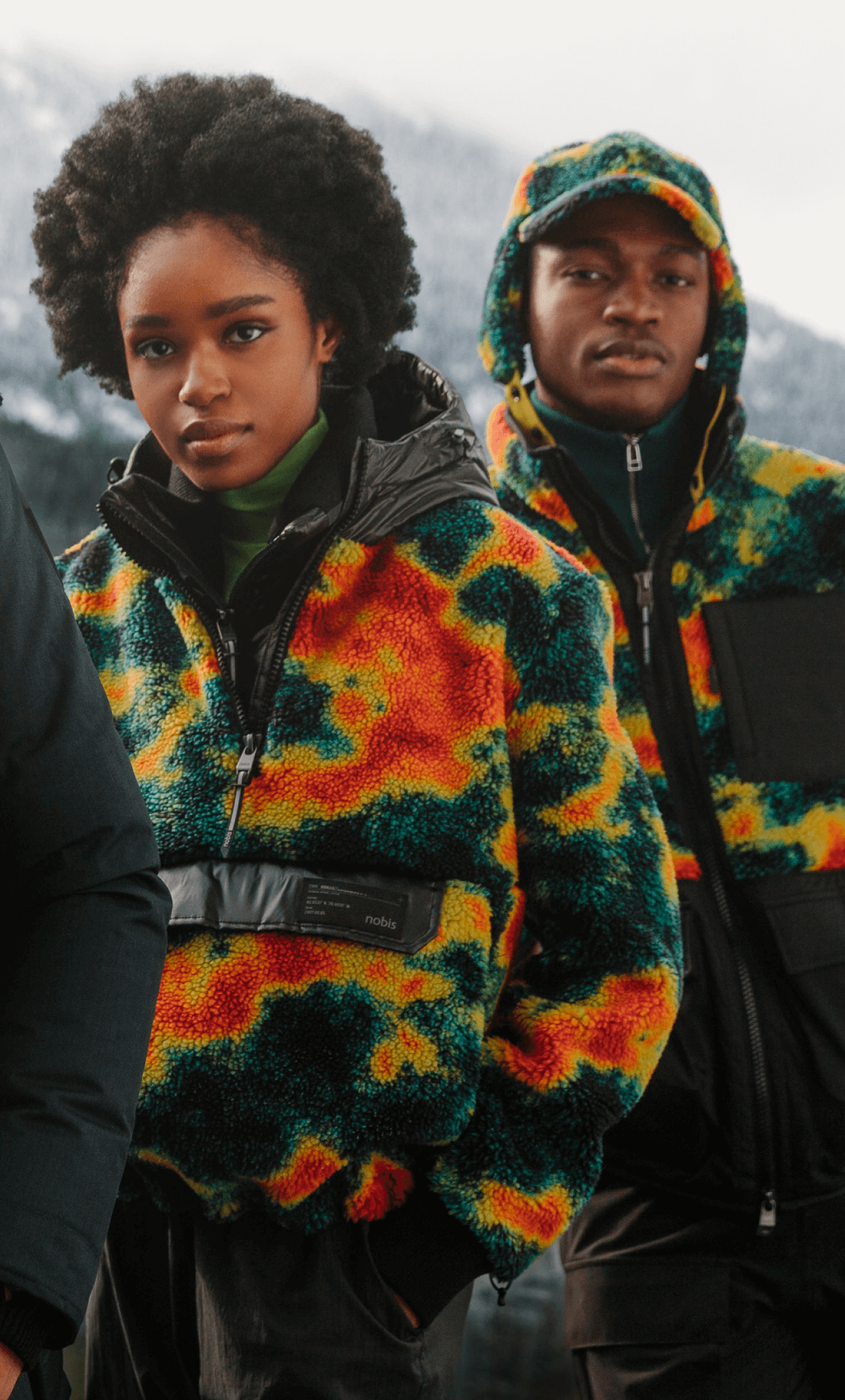 Two models lean on a railing with snowy mountains in the background. Woman on left wears Nobis Roche in Heat Map. Man on right wears Nobis Kepler in Heat Map.